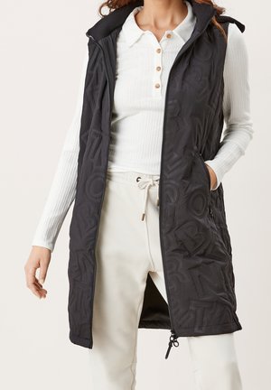 Woman wearing long black quilted sleeveless vest over white ribbed long-sleeve shirt and cream drawstring pants, hand in vest pocket.