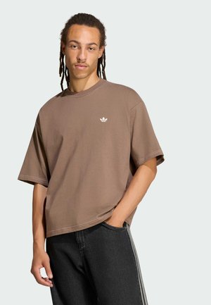 Junger Mann mit Dreadlocks, der ein lockeres braunes T-Shirt mit kleinem weißem Logo und schwarze Hose mit weißen Streifen trägt, posiert mit einer Hand in der Tasche.
