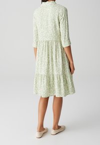 Robe verte à motifs avec une jupe à volants et des manches trois-quarts, fabriquée en tissu doux. Portée avec des chaussures légères à enfiler.