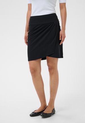 Femme portant une jupe portefeuille asymétrique noire, un top blanc et des ballerines noires, debout devant un fond clair uni.