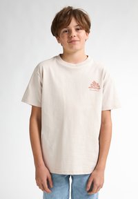 Beige katoenen t-shirt met korte mouwen, ronde hals, met een roze grafisch ontwerp op de linkerkant van de borst. Draagt met lichtblauwe jeans.