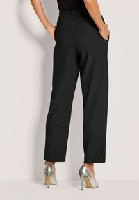 Vrouw in gestructureerde zwarte wijde broek en zilveren hoge hakken, staand met handen in de zakken tegen een lichte muur.