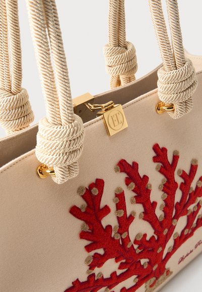 Μπεζ τσάντα tote με χοντρές λαβές από σχοινί που είναι δεμένες σε χρυσά κρίκους, χρυσό κλείστρο και κόκκινη κεντητή κοραλλιογενής διακόσμηση στην εξωτερική πλευρά του υφάσματος.