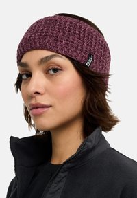 Knit, maroon headband featuring a textured pattern and a logo patch. Worn with a black fleece jacket. Close-up view of the wearer’s face.
