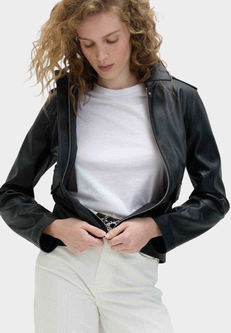 Woman with curly hair wearing black leather jacket over white shirt and light pants, fastening the jacket zipper.