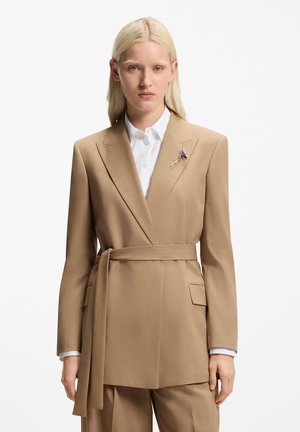 Bézs szabott blézer átkötős résszel, övvel, kivágott hajtókával és zsebbel. Fehér inggel, kék részletekkel viselve.