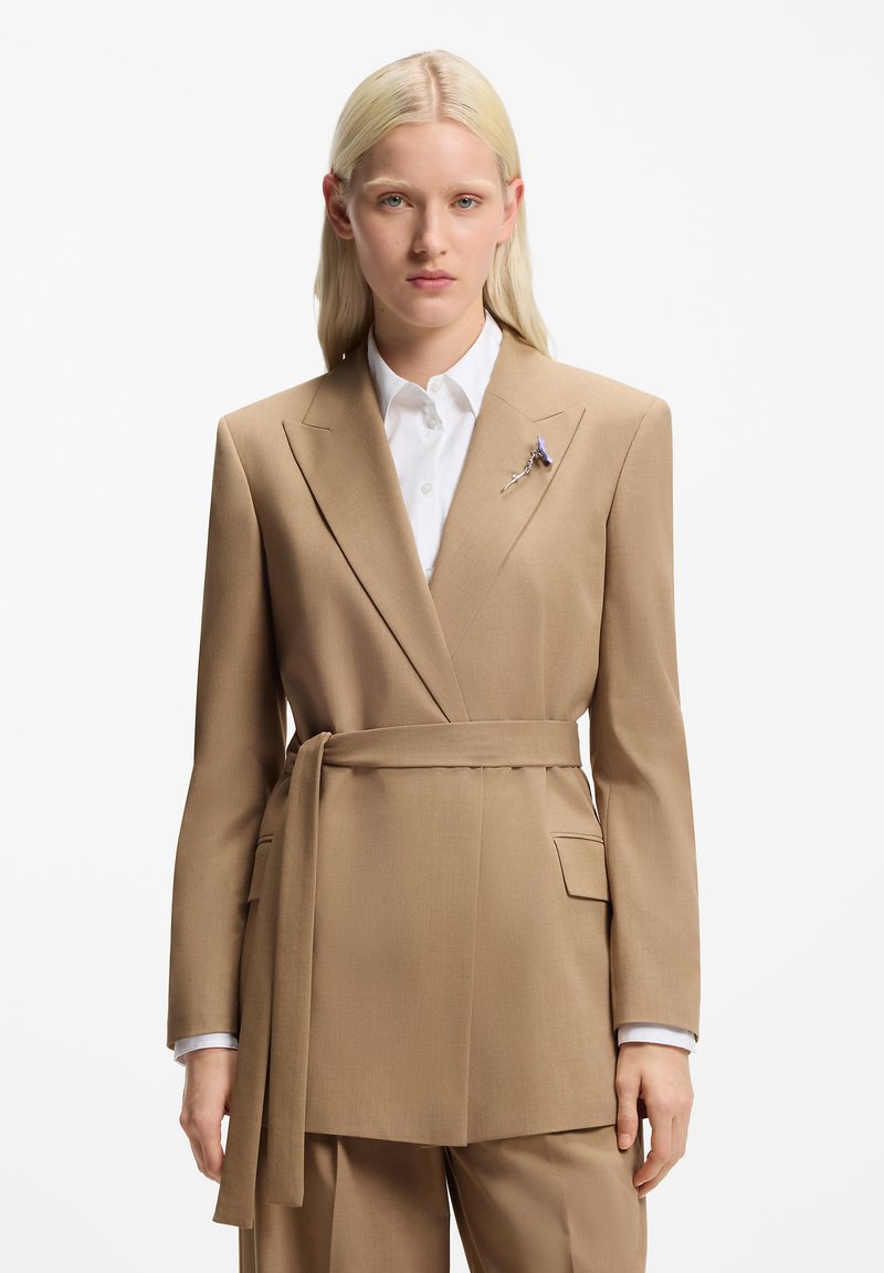 Μπεζ σακάκι ραμμένο στο μέτρο με σχεδιασμό wrap, λεπτομέρειες ζώνης, πέτο σε σχήμα υποδοχής και τσέπη. Φοριέται πάνω από λευκό πουκάμισο με μπλε λεπτομέρειες.