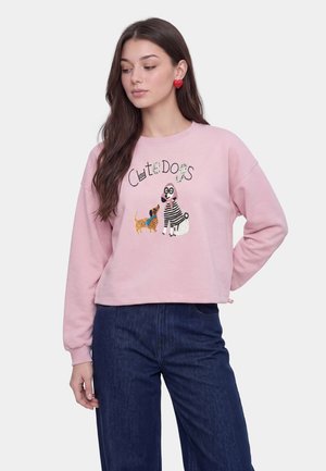 Vrouw draagt een roze sweatshirt met geborduurde honden en tekst "Cute Dogs", gecombineerd met donkerblauwe spijkerbroek en rode oorbellen in hartvorm.