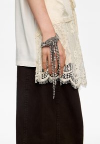 Ręka nosząca srebrną bransoletkę z kryształkami rhinestone, z zawieszonymi nitkami, na tle kremowej koronki i ciemnobrązowej spódnicy.