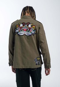 Khaki kurtka z dużym haftowanym wzorem orła na plecach oraz różnymi naszywkami na rękawach. Teksturowany materiał, kroj militarny.