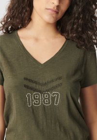 Vrouw in een olijfgroene V-hals t-shirt met zwarte chevronstrepen en witte geborduurde "1987" aan de voorkant.
