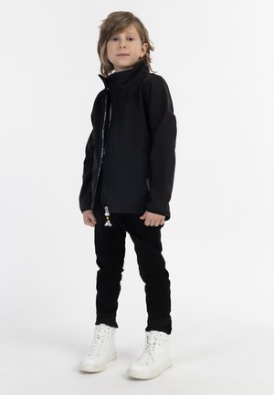 Kind mit hellbraunen Haaren steht nach vorne gerichtet, trägt eine schwarze Jacke, schwarze Hose und weiße High-Top-Sneaker vor weißem Hintergrund.