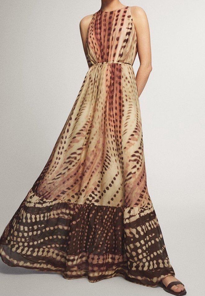 Robe longue fluide sans manches avec un motif tacheté abstrait dans des tons beige, marron et rouges, portée avec des sandales noires.
