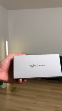 Grijze rechthoekige doos met afgedrukte logo's: "Ray-Ban" en "Meta." Gladde textuur, vastgehouden in een open handpalm, met een wazige achtergrond.