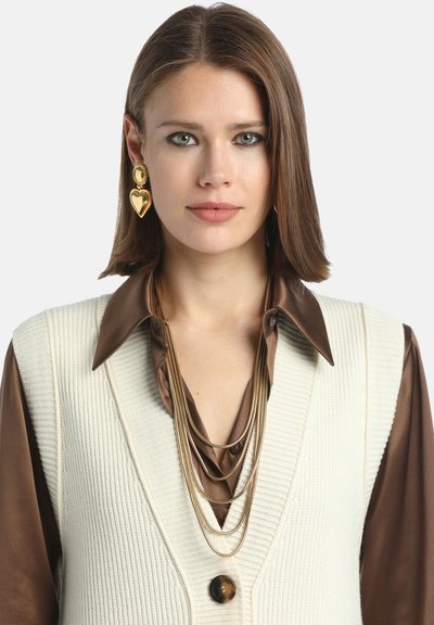 Auksinė pakabukas su sluoksniais ant rudos bluzonos ir kreminės megztos liemenės su dideliu mygtuku, kairėje ausyje - širdelės formos auskaras.