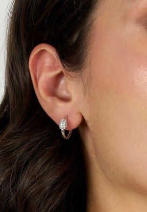 Orecchini a cerchio in argento con un grappolo circolare di piccoli gemelli trasparenti. In primo piano l'orecchio con capelli scuri visibili.