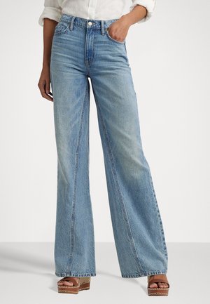 Vrouw die lichtblauwe, hoog getailleerde, wijd uitlopende jeans draagt, een wit overhemd met opgerolde mouwen en bruine platformsandalen, tegen een effen achtergrond.