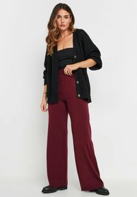 Svart stickad cardigan med stora knappar, i kombination med en åtsittande svart topp och vida bourgognebyxor. Modellen bär svarta ankelstövlar.