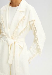 Bata de color crema con acentos de borlas en las mangas, un cinturón atado a la cintura y un cuello. Hecha de una tela texturizada.