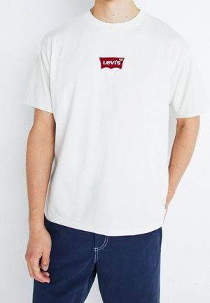 Mężczyzna ubrany w białą koszulkę Levi's z czerwonym logo na piersi oraz granatowe spodnie, stojący na białym tle.