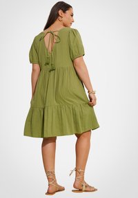 Olivgrünes, knielanges Kleid mit kurzen Puffärmeln, gestuften Rüschensaum und einer Schleife am Rücken mit Quasten. Getragen mit beigen Sandalen.