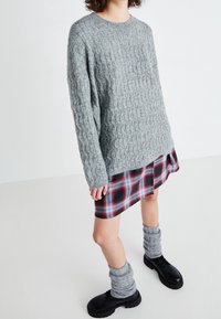 Person, die einen grauen Zopfmuster-Pullover, einen Karo-Rock, graue Stulpen und schwarze Schnürschuhe trägt, vor einem schlichten weißen Hintergrund.