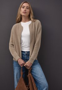 Cardigan beige duveteux avec une fermeture éclair à l'avant, porté sur une chemise blanche. Un jean bleu et un sac en daim marron complètent la tenue. Le fond est gris foncé.