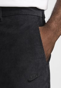 Svarta kordflamsbyxor med vertikala ribbor, med en sidoficka och en svart Nike-logotyp broderad på framsidan.