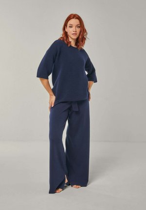 Young woman with red hair wearing loose navy blue top and wide-leg pants standing barefoot against plain gray background.