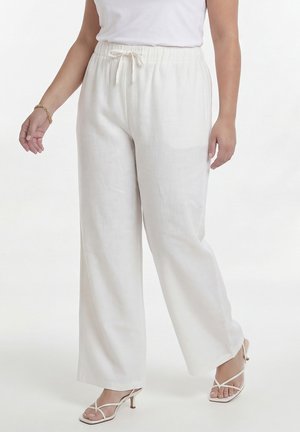 Kvinde iført løstsiddende hvide hørbukser med elastisk talje og snøre, kombineret med hvide højhælede sandaler med stropper.
