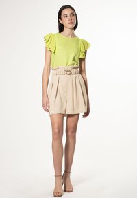 Limegrøn top med volangærmelige i blonde, parret med beige plisserede shorts med samlet talje og rund guldspænde.