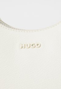 Balta odinė rankinė su tekstūruota paviršiumi ir iškilmingu "HUGO" logotipu šviesioje spalvoje arti viršaus, su lygaus užtrauktuko detale.