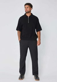 Pull noir à manches courtes et zip quart, texture tricotée, associé à un pantalon noir et des chaussures noires avec une semelle beige, créant un look monochrome.