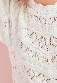 Pletený svetr v barvě off-white s jemnými krajkovými vzory, strukturovaným povrchem a zvýšeným límcem. Obsahuje detaily s otevřenými vzory a volný střih.