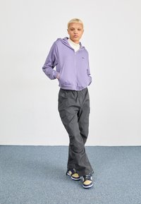 Felpa con zip viola, pantaloni cargo grigi e sneakers multicolori. Tessuto morbido, vestibilità comoda e dettagli di cucitura visibili sui pantaloni.