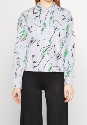 Femme portant une chemise blanche à boutons avec des motifs abstraits de lignes vertes, bleues et noires, associée à un pantalon noir à jambes larges.