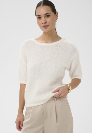 Woman wearing a white fuzzy short-sleeve sweater with beige pleated pants, gold hoop earrings, and a chunky gold bracelet.