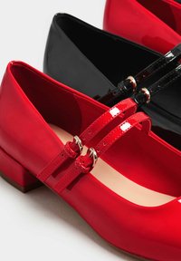 Punaiset ja mustat kiiltävät Mary Jane -kengät, joissa on pyöreä kärki, kaksi ohutta remmiä ja pienet metalliset soljet. Matalan lohko-kannan muotoilu.