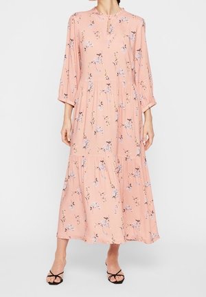 Mujer con un vestido rosa de manga larga, hasta el tobillo y con estampado floral, y sandalias negras de tiras, de pie contra un fondo blanco liso.