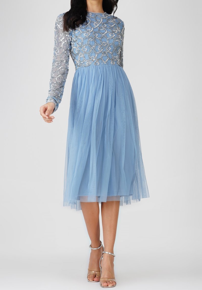 Femme portant une robe midi bleu clair avec corsage perlé argenté et manches longues transparentes, associée à des sandales argentées à talons hauts.
