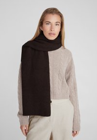 Écharpe côtelée marron foncé au toucher doux, drapée autour du cou, associée à un pull en maille torsadée beige clair.