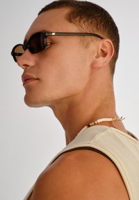 Άνδρας που φορά στενά μαύρα γυαλιά ηλίου, εκρού αμάνικη μπλούζα και κολιέ με χάντρες, κοιτάζοντας προς τα αριστερά με ουδέτερη έκφραση.