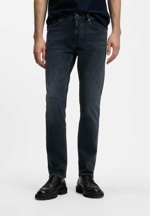 Mand iført mørkeblå slim-fit jeans og sorte lædersko står foran en ensfarvet baggrund.