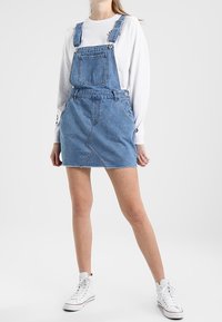 Denimoverallklänning med justerbara axelremmar, ficka fram och silverknappdetaljer. Har en ljusblå färg och en åtsittande kjol.