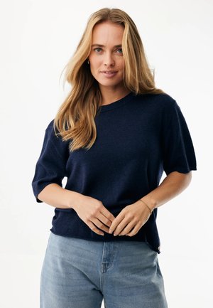 Blonde Frau trägt einen marineblauen kurzärmligen Pullover und hellblaue Jeans, steht mit vor dem Körper verschränkten Händen vor einem weißen Hintergrund.