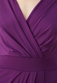 Violetinė suknelė su giliu V formos iškirpimu, dekoruota drapiruotėmis ir prigludusia juosmens dalimi. Pagaminta iš minkštos medžiagos su glotna tekstūra.