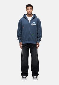 Hooded zip-up sweatshirt in dark blue, featuring white text and colored embroidered patches. Paired with loose black jeans and black sneakers.
