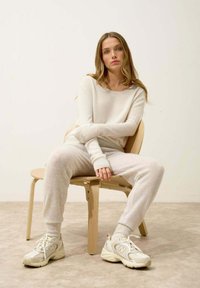 Maglione a maglia color crema e pantaloni jogger coordinati, abbinati a sneakers chiare. L'ambientazione presenta una sedia in legno minimalista.