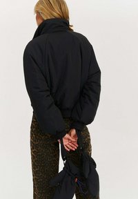 Čierna prešívaná bunda s vysokým golierom a stiahnutými manžetami, nosená na nohaviciach so vzorom hnedých leopardích škvŕn. Drží čierne odnímateľné rukavice.