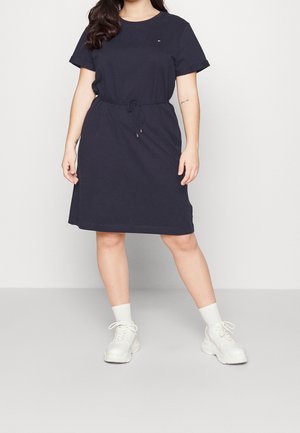 Kobieta stoi, ubrana w granatową sukienkę z krótkim rękawem, z paskiem ściągającym w talii oraz białe trampki z białymi skarpetkami.